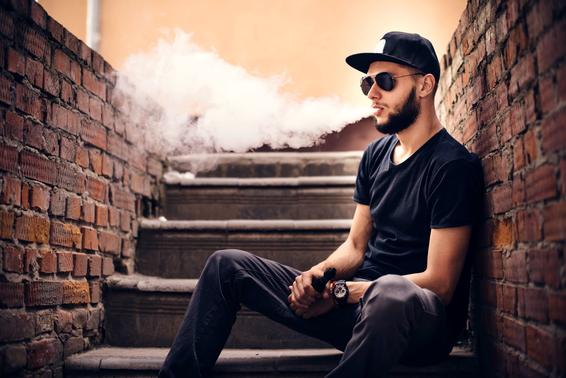 Man in sunglasses and hat exhaling vape sitting on brick steps